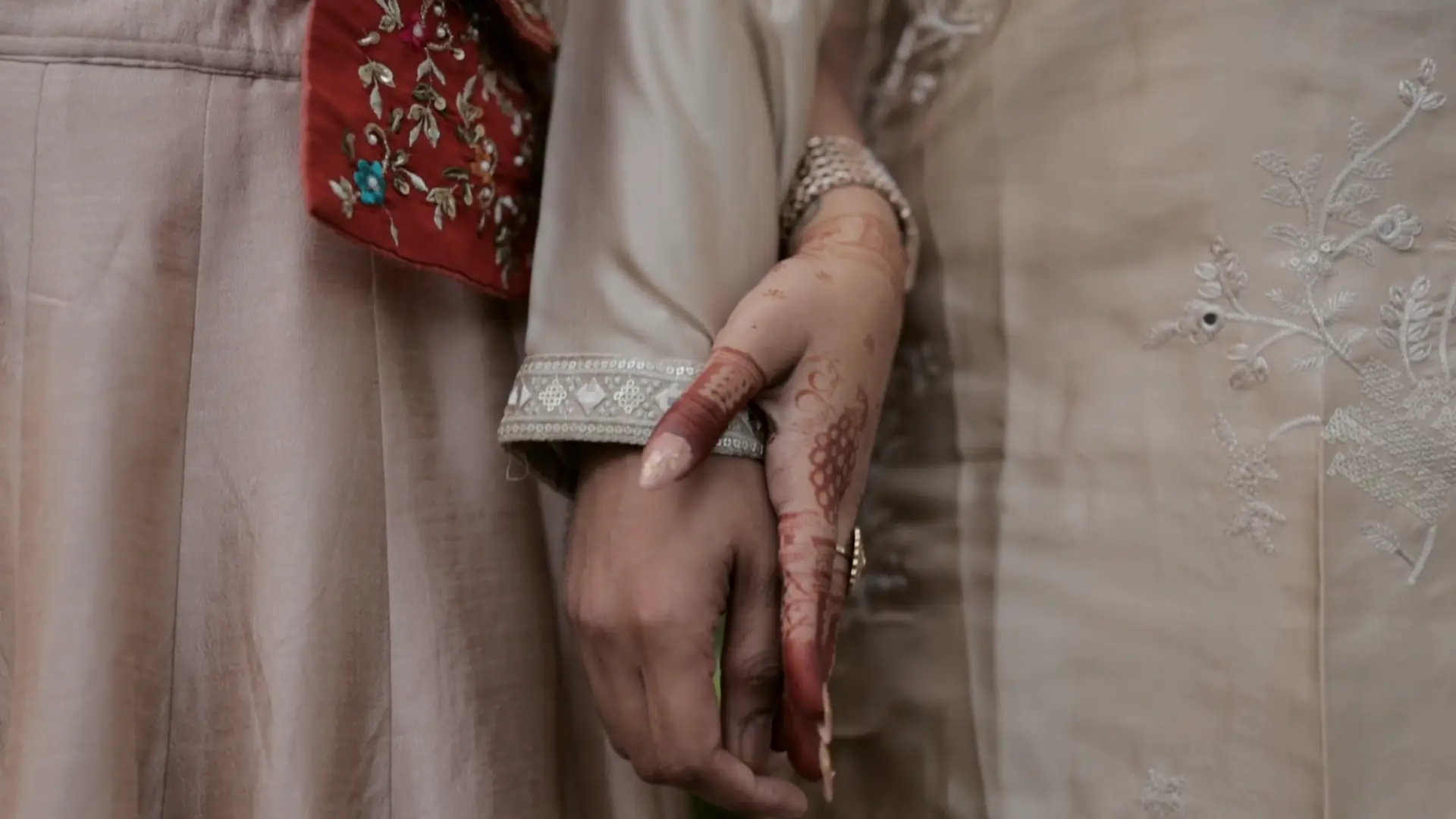 The bride's hennaed hand holding the groom's against embroidered clothes