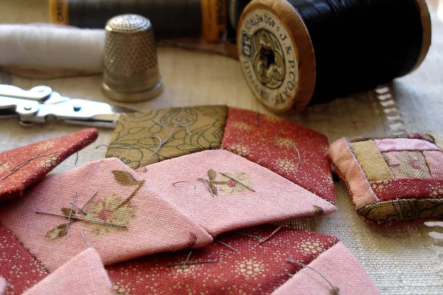 Pink and olive fabric patches, a thimble and spools of black thread on a table, a baby's quilt half stitched