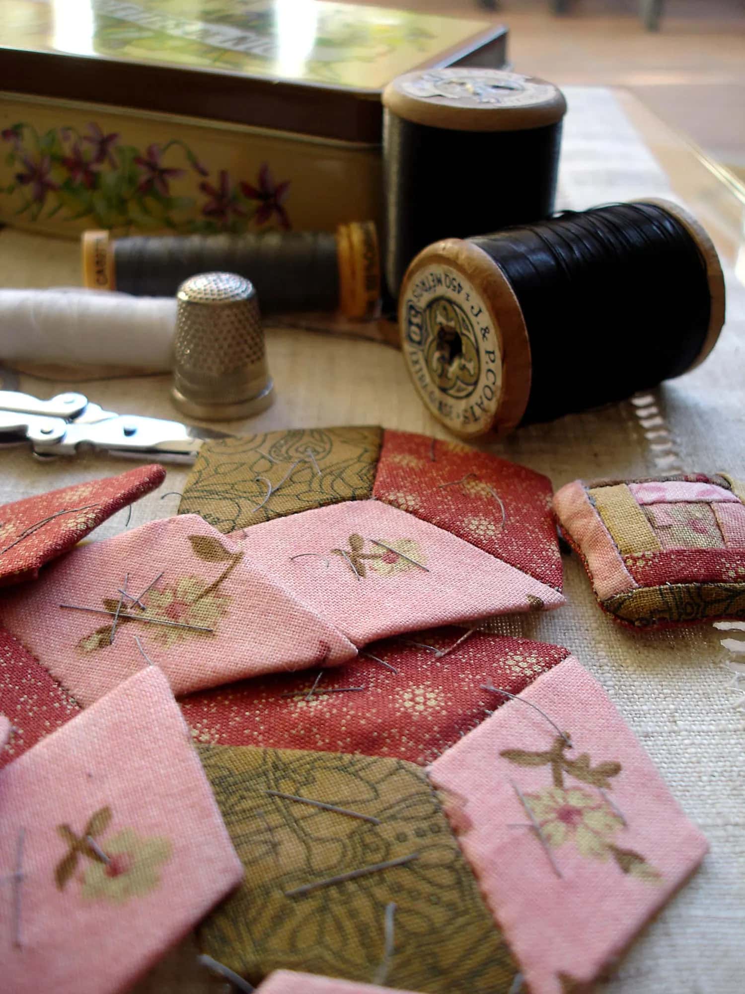 Pink and olive fabric patches, a thimble and spools of black thread on a table, a quilt half stitched