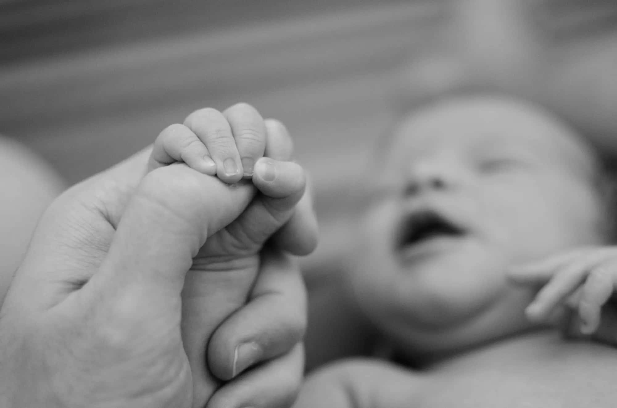 A newborn's hand holding tightly to a parent's finger, in black and white