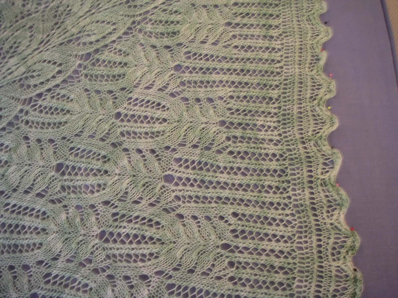 A pale green hand-knitted lace baby blanket pinned out flat