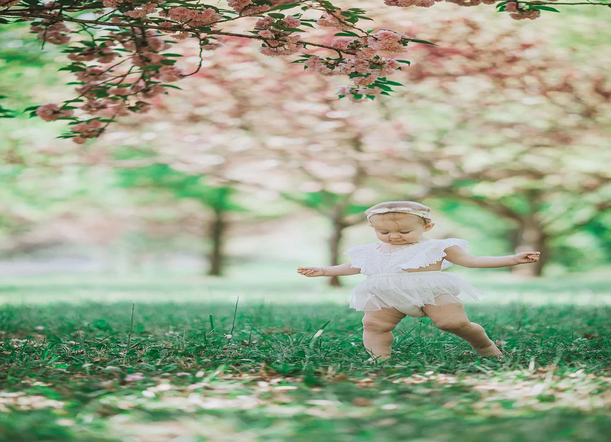 A toddler's bare legs and one hand in the grass mid-step, her white frock swinging