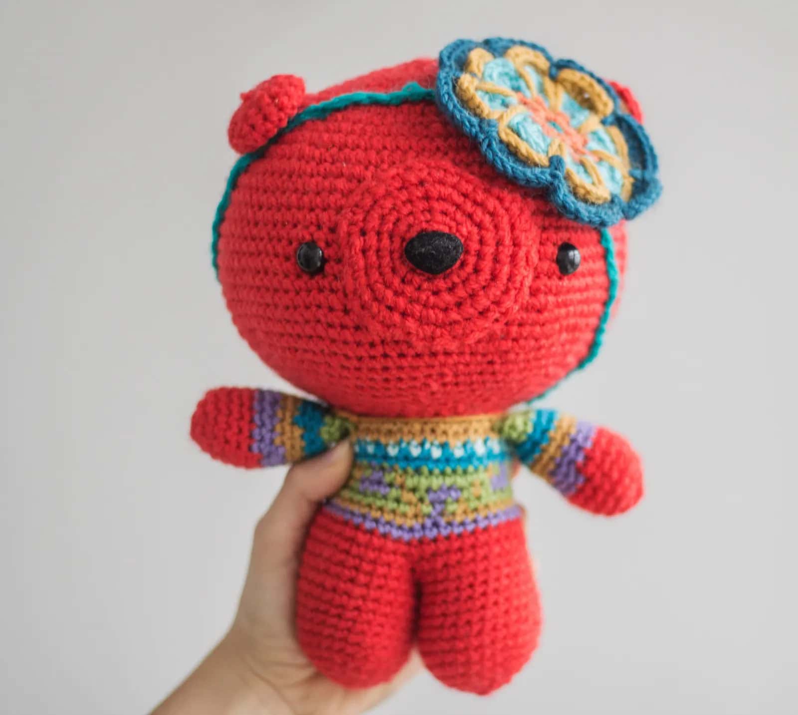 A hand holding a crocheted red teddy bear in a striped jumper with a flower on its ear