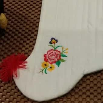 A detail of an embroidered flower on a white baby sock, a red tassel beside it