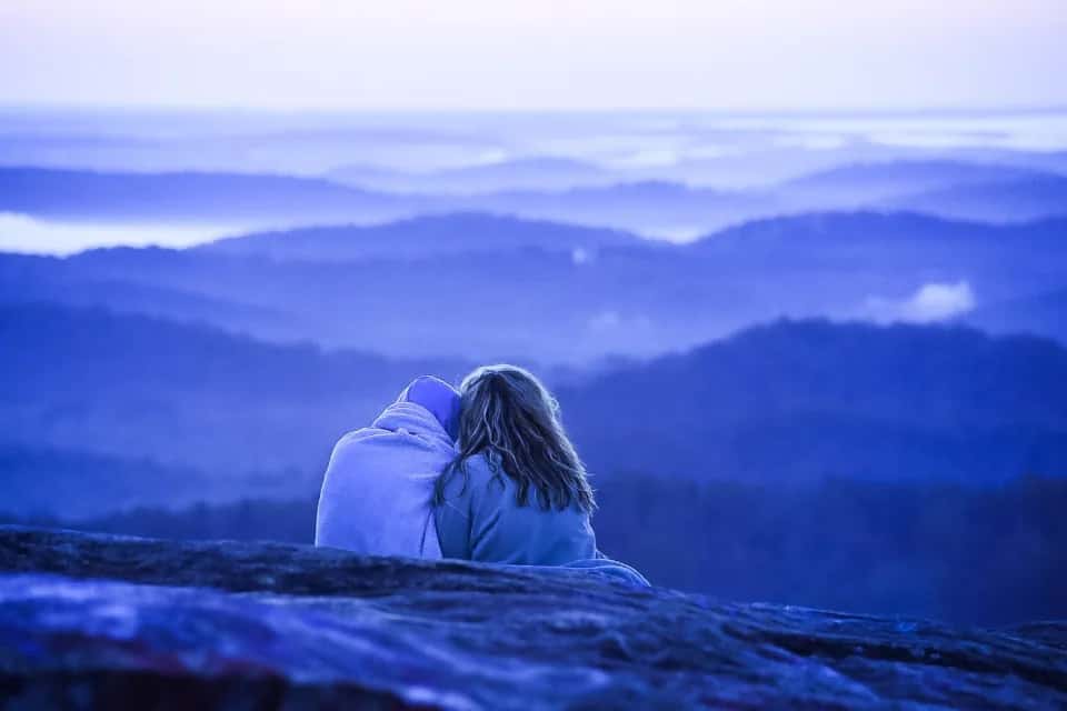 A couple sitting close together, looking out over hills folded in blue mist