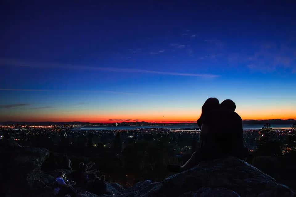 A couple in silhouette on a rock above a city's lights at dusk, the sky orange at the horizon