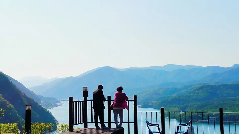 A couple seen from behind at a railing, looking out over a lake between misty hills