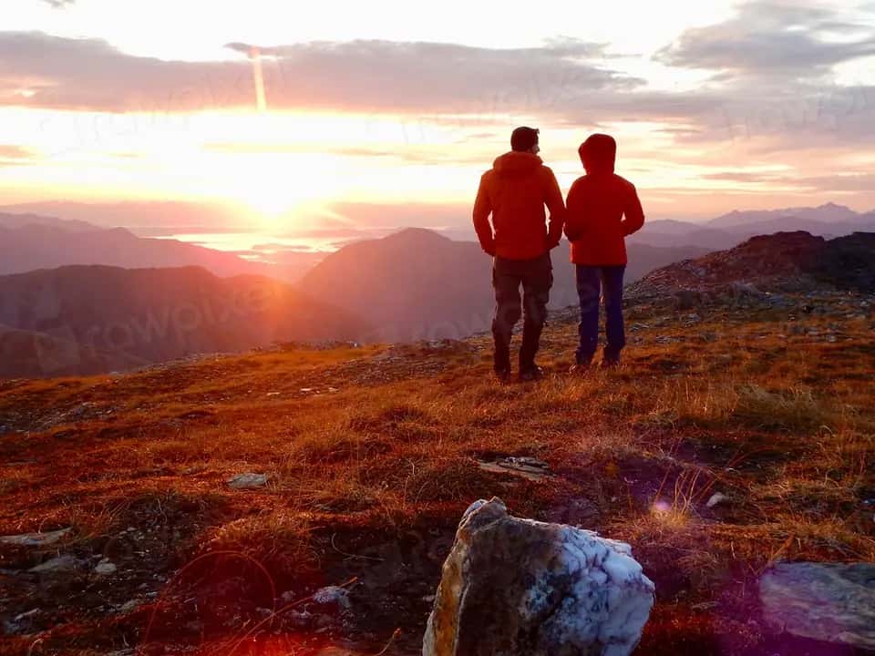 A couple in red jackets, seen from behind, watching the sun set over a range of mountains