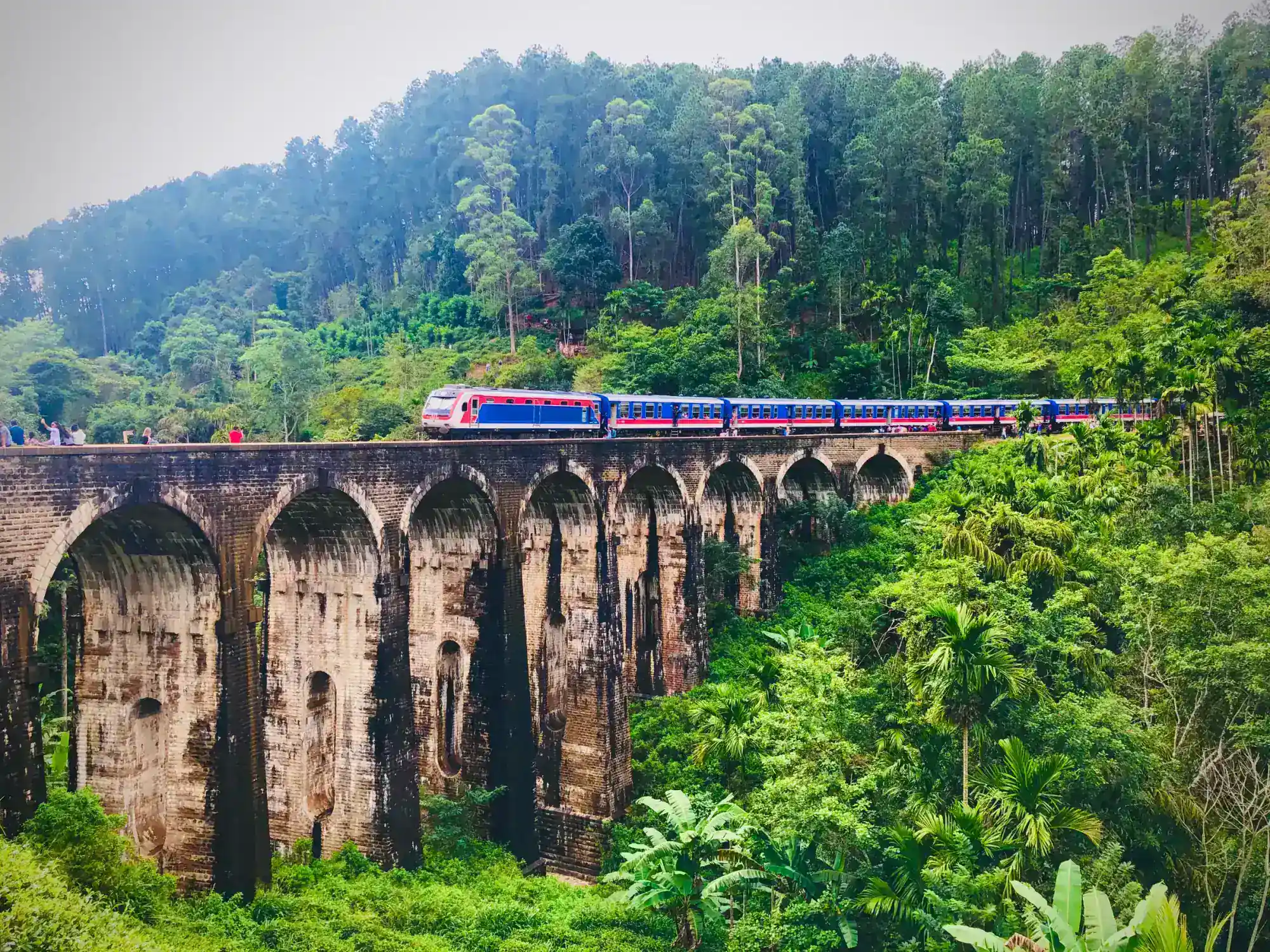 A blue and white train crossing the Nine Arch Bridge among green hills