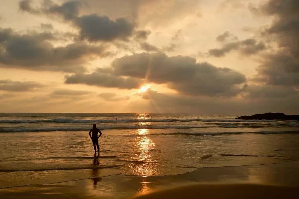 One person standing in the shallows at Bentota as the sun sets gold through the clouds