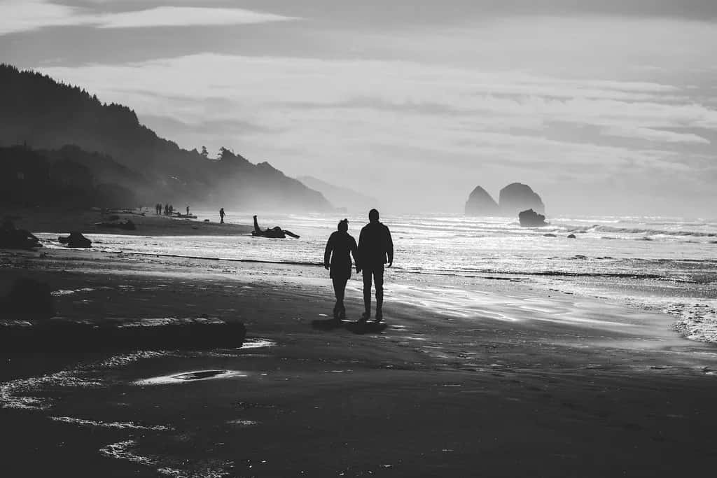 A couple walking hand in hand along a grey shore, sea stacks in the haze behind them