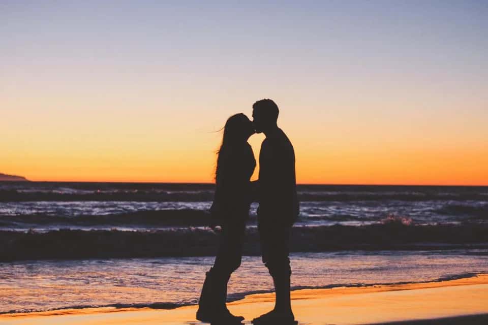 A couple in silhouette on a beach at sunset, the waves orange behind them