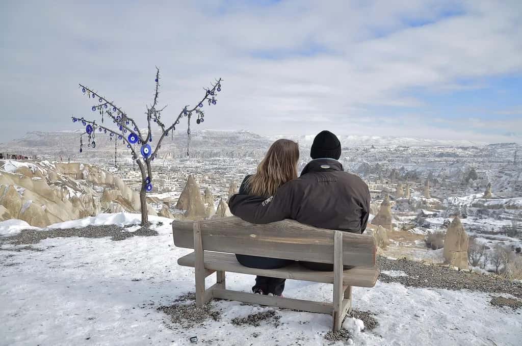 A couple seen from behind on a bench above the snowy rock valleys of Cappadocia, an evil-eye tree beside them