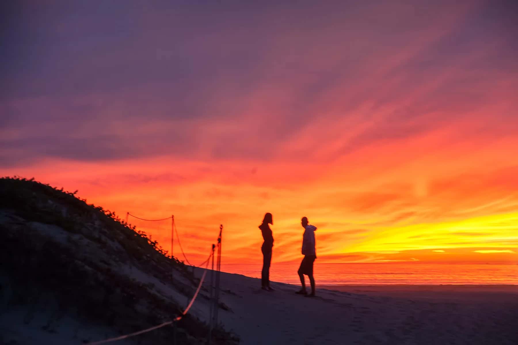 Two small figures on a sand dune against a blazing orange sunset