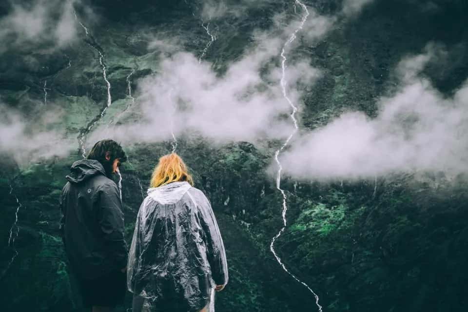 Two people in rain jackets, seen from behind, facing misty green mountains