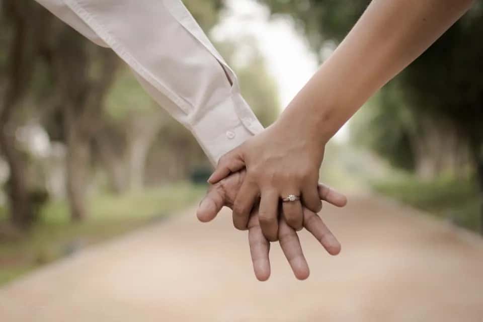 Two hands holding on a tree-lined path, a ring on hers