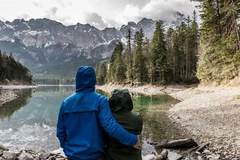 A couple seen from behind, arm in arm at a clear lake below pine forest and peaks