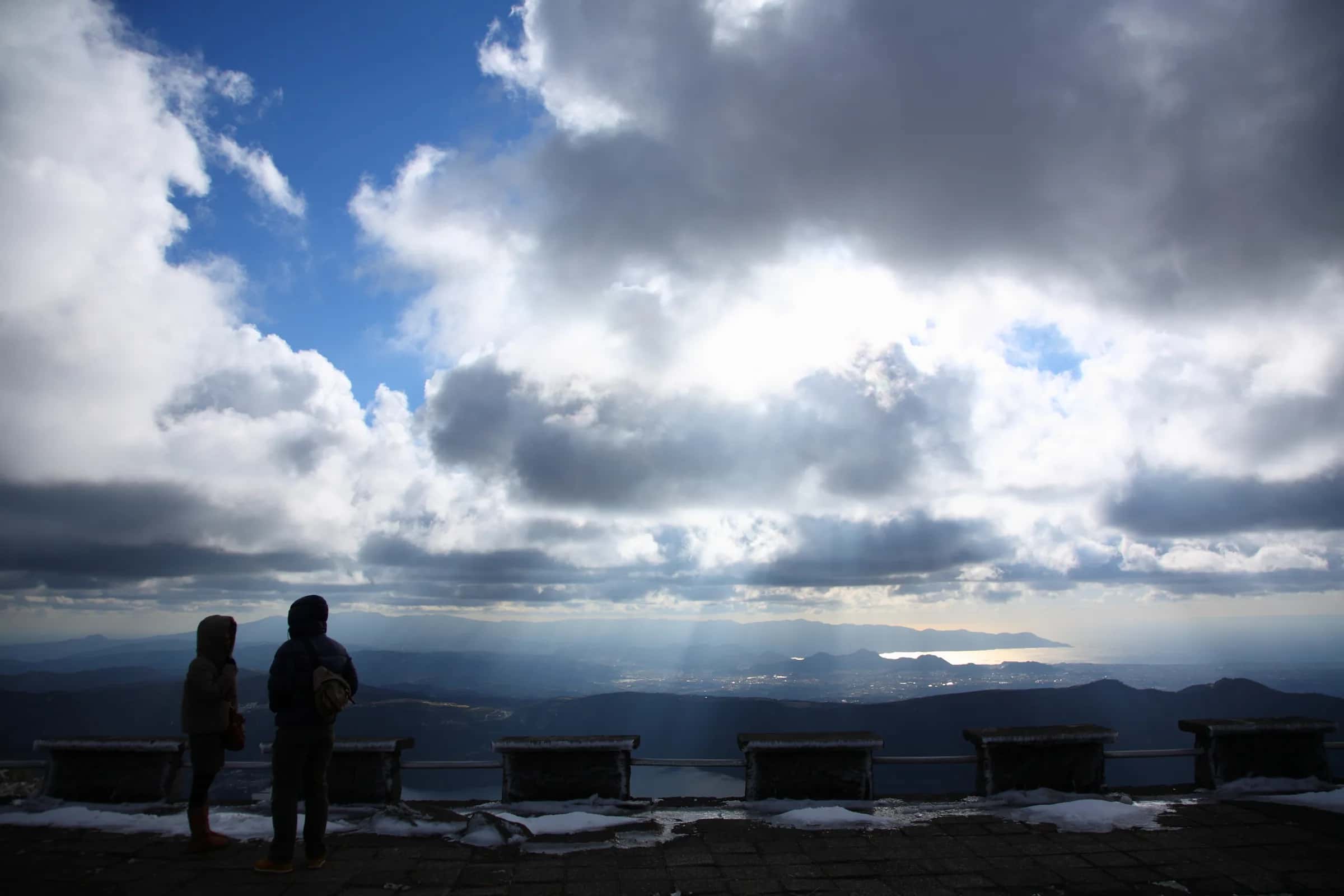 Two people in winter coats at a snowy lookout, looking out over the valleys under breaking cloud