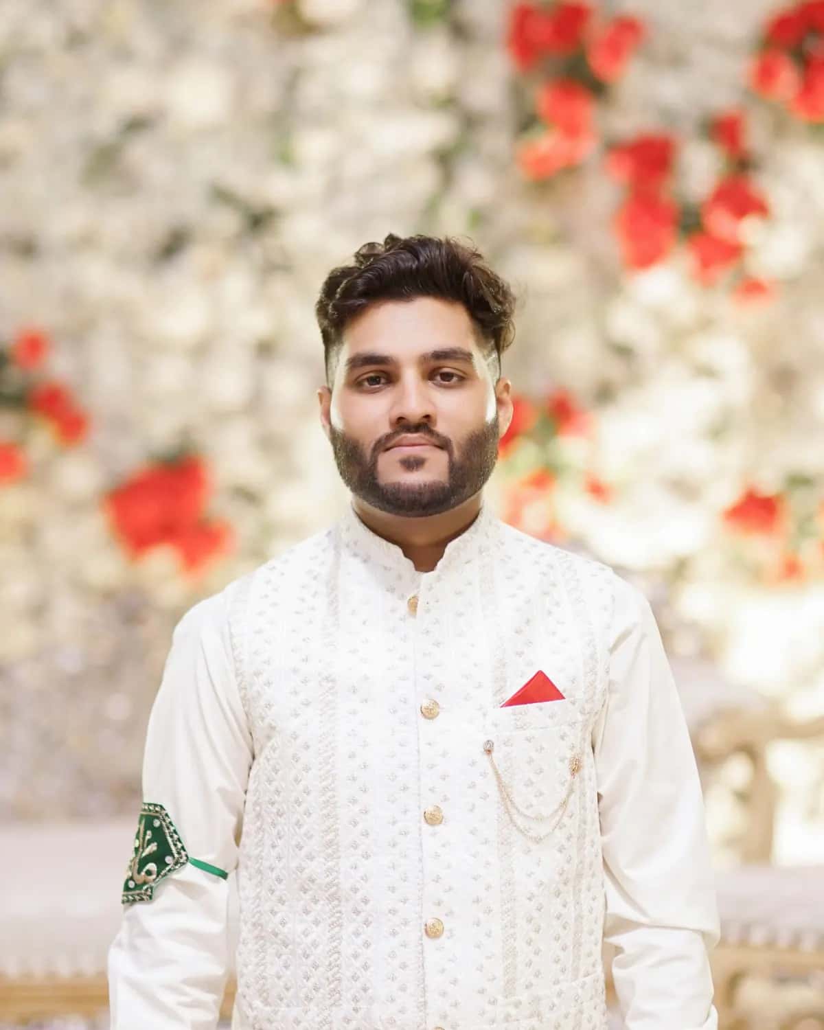 The groom in a white sherwani before a wall of flowers