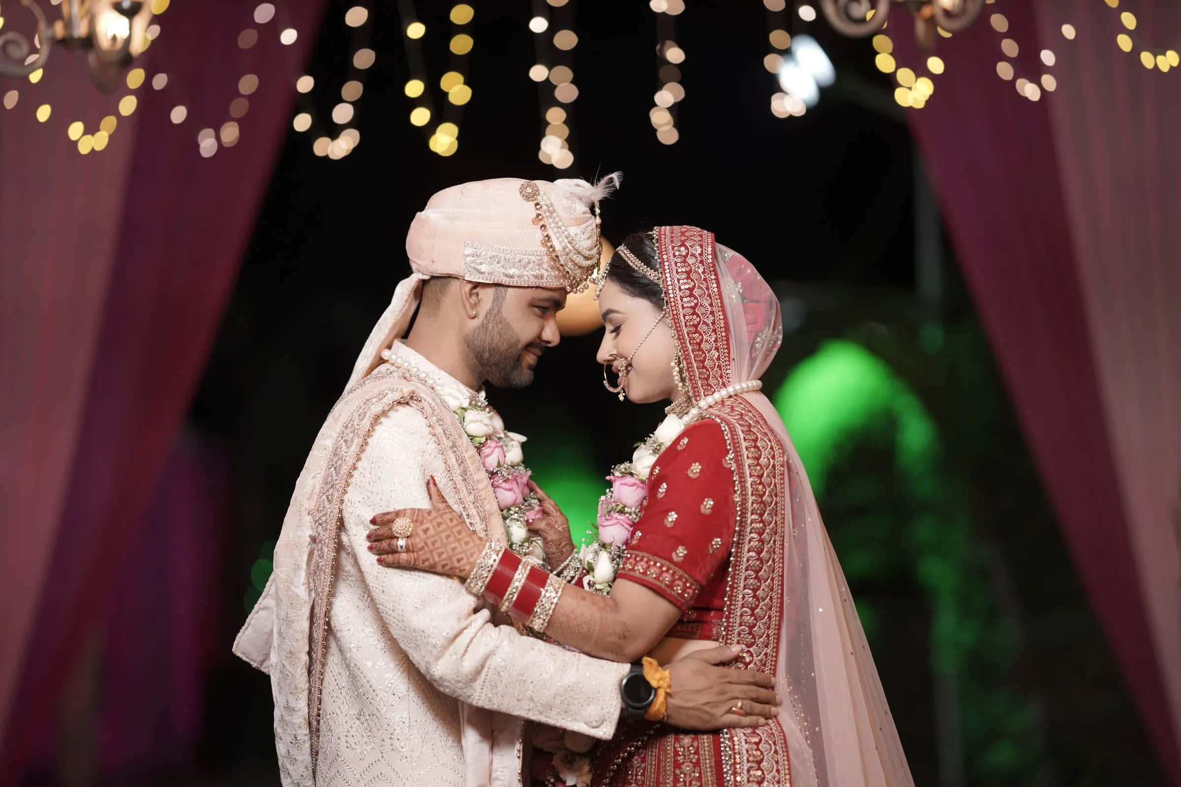 A bride and groom holding each other under fairy lights and deep red drapes