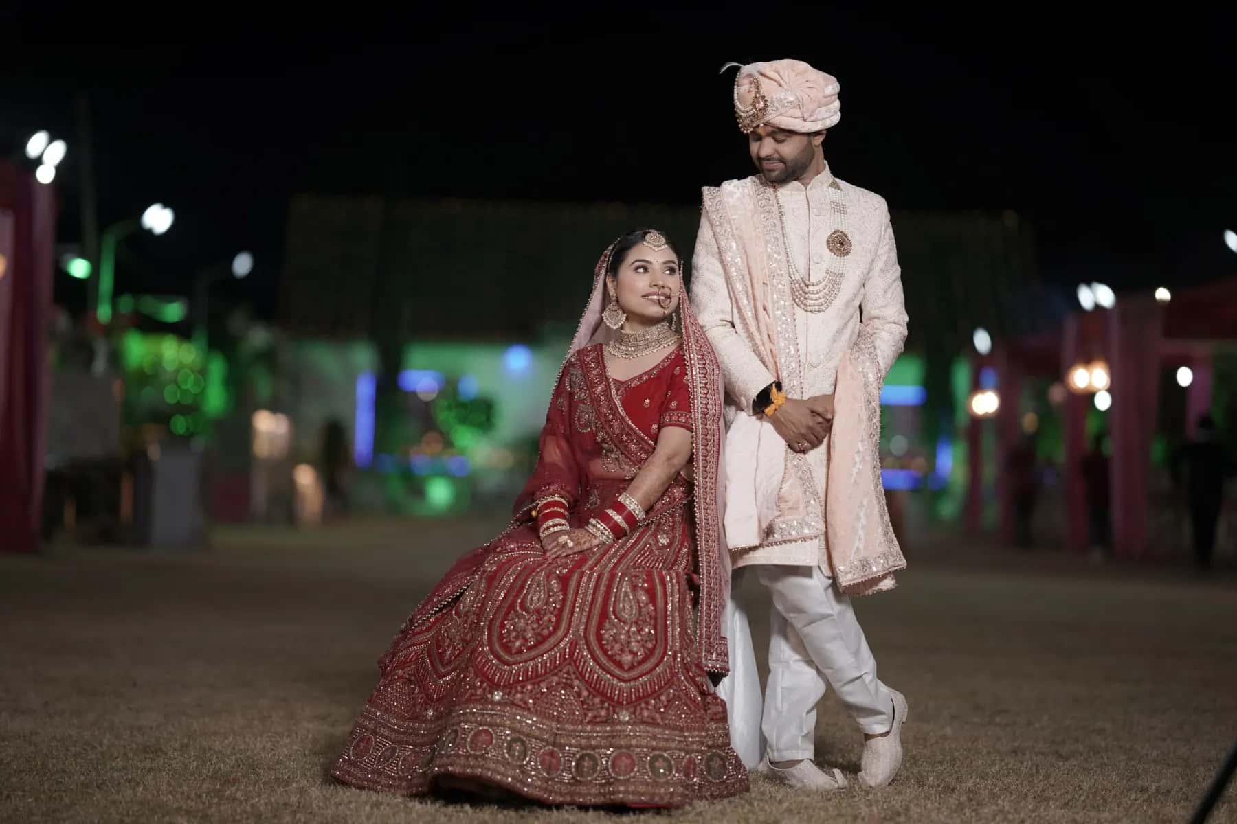 The bride in red seated beside the groom in an ivory sherwani, the lights of the marquee behind them