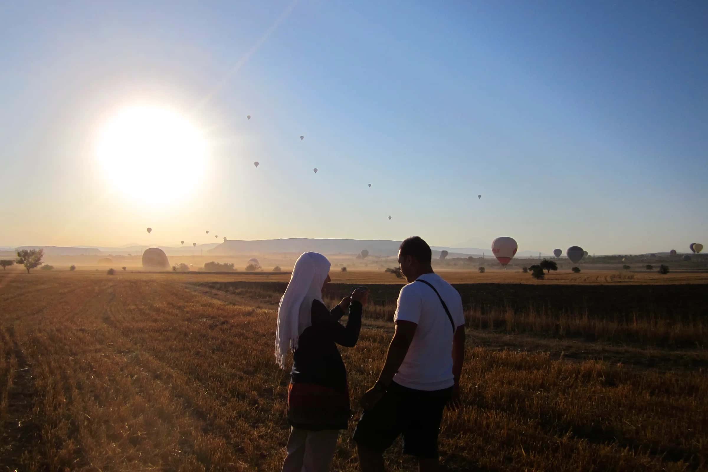 A couple seen from behind in a field at sunrise, she in a white scarf, hot-air balloons rising over the plain beyond
