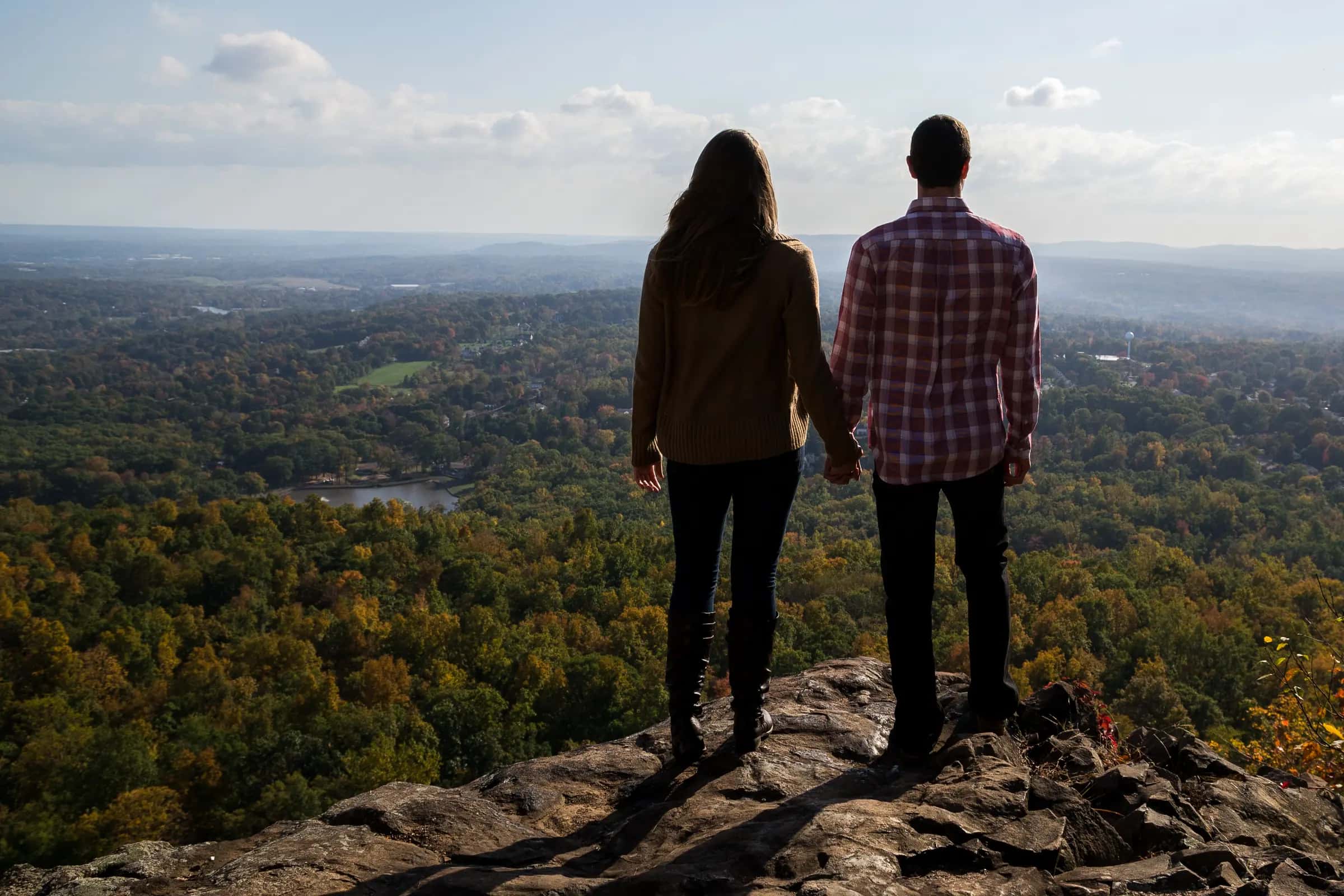 A couple seen from behind, hand in hand on a rock ledge, looking out over an autumn valley